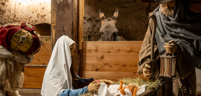 Das Bild zeigt eine Weihnachtskrippe im Heimatmuseum Sixenhof in Achenkirch mit Figuren von Maria, Josef und einem Neugeborenen im Stroh. Im Hintergrund ist ein Esel zu sehen.
