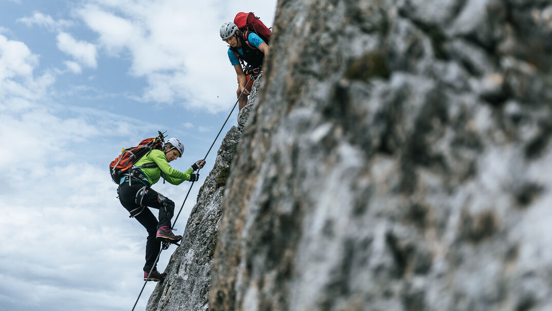 Ein Mann und eine Frau auf einem Klettersteig im Rofangebirge bei wenig bewölktem Himmel.