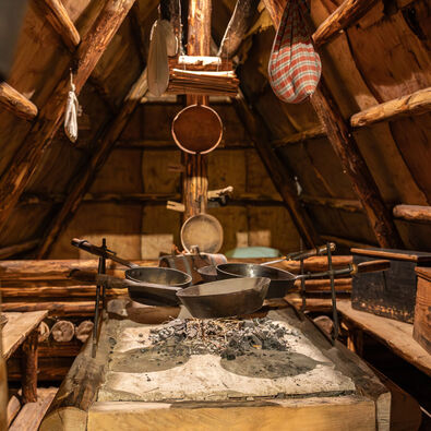 An interior view of the rustic kitchen of the Local history museum Sixenhof, showcasing a stone cooking area with several black pans suspended above a small fire made of charcoal.