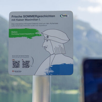 Ein Informationsschild am Aussichtspunkt Hochsteg am Achensee. Es bewirbt eine interaktive Hörspiel-Tour mit Kaiser Maximilian I. und enthält Anweisungen zum Scannen eines QR-Codes, um mehr über die Geschichte und die Umgebung zu erfahren.