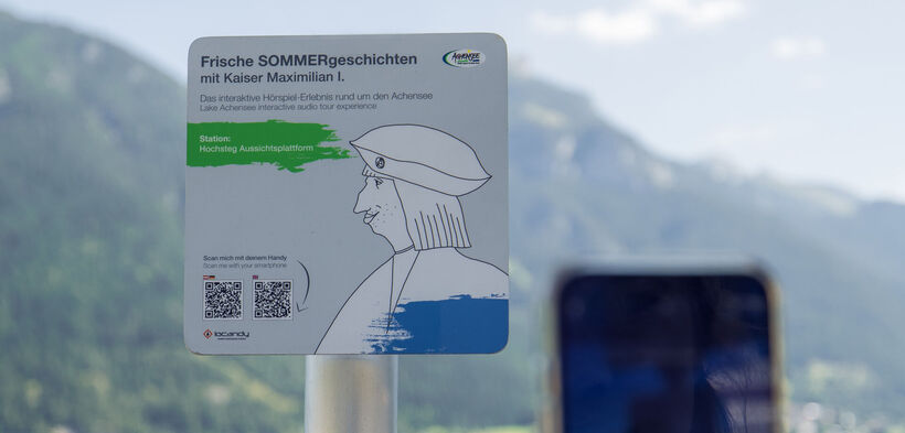 A sign promoting an interactive audio experience titled "Fresh Summer Stories with Emperor Maximilian I." It indicates the location as "Hochsteg Viewing Platform" and includes instructions to scan a QR code with a smartphone for more information. The background features mountains and a lake.
