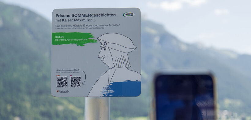 A sign promoting an interactive audio experience titled "Fresh Summer Stories with Emperor Maximilian I." It indicates the location as "Hochsteg Viewing Platform" and includes instructions to scan a QR code with a smartphone for more information. The background features mountains and a lake.