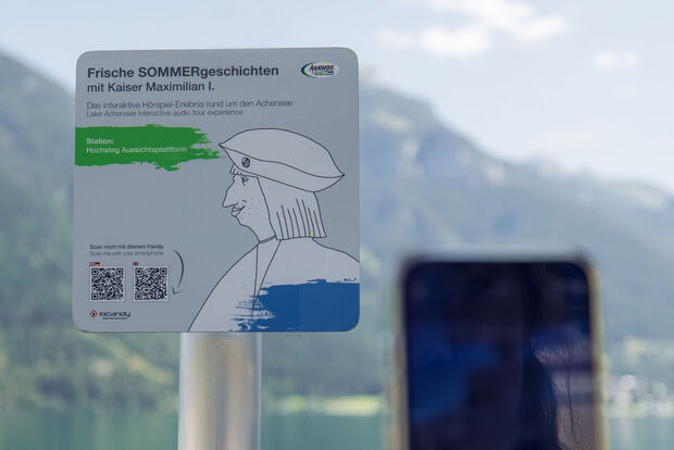 A sign promoting an interactive audio experience titled "Fresh Summer Stories with Emperor Maximilian I." It indicates the location as "Hochsteg Viewing Platform" and includes instructions to scan a QR code with a smartphone for more information. The background features mountains and a lake.