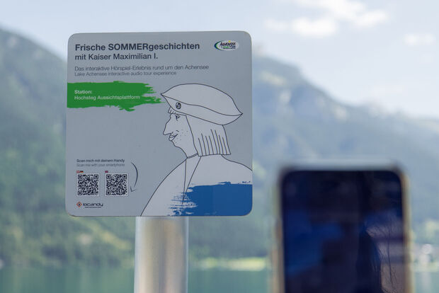 A sign promoting an interactive audio experience titled "Fresh Summer Stories with Emperor Maximilian I." It indicates the location as "Hochsteg Viewing Platform" and includes instructions to scan a QR code with a smartphone for more information. The background features mountains and a lake.