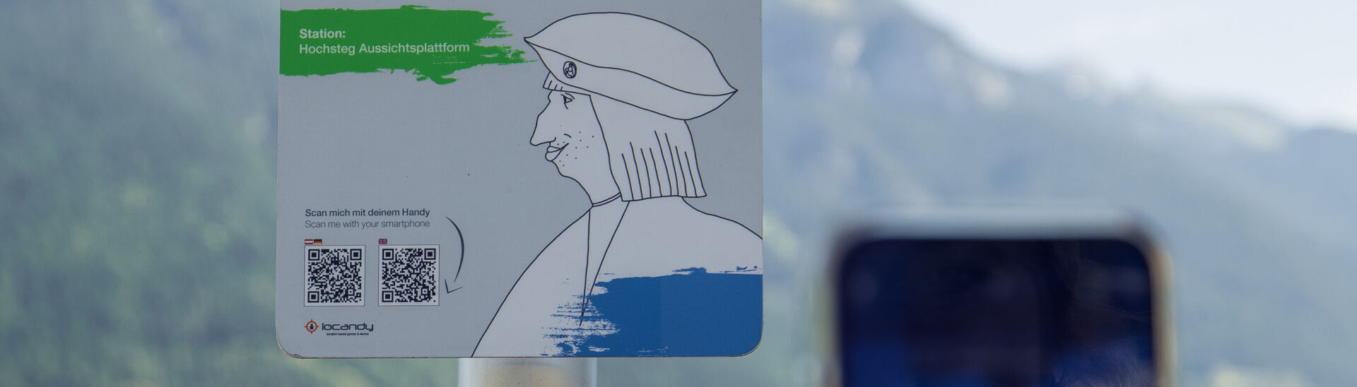 Hörspiel-Erlebnis am Achensee mit Kaiser Maximilian I. Ein Informationsschild am Aussichtspunkt Hochsteg am Achensee. Es bewirbt eine interaktive Hörspiel-Tour mit Kaiser Maximilian I. und enthält Anweisungen zum Scannen eines QR-Codes, um mehr über die Geschichte und die Umgebung zu erfahren.