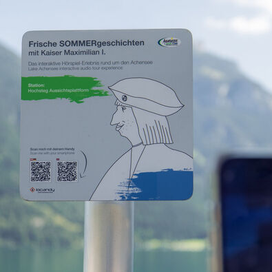 A sign promoting an interactive audio experience titled "Fresh Summer Stories with Emperor Maximilian I." It indicates the location as "Hochsteg Viewing Platform" and includes instructions to scan a QR code with a smartphone for more information. The background features mountains and a lake.
