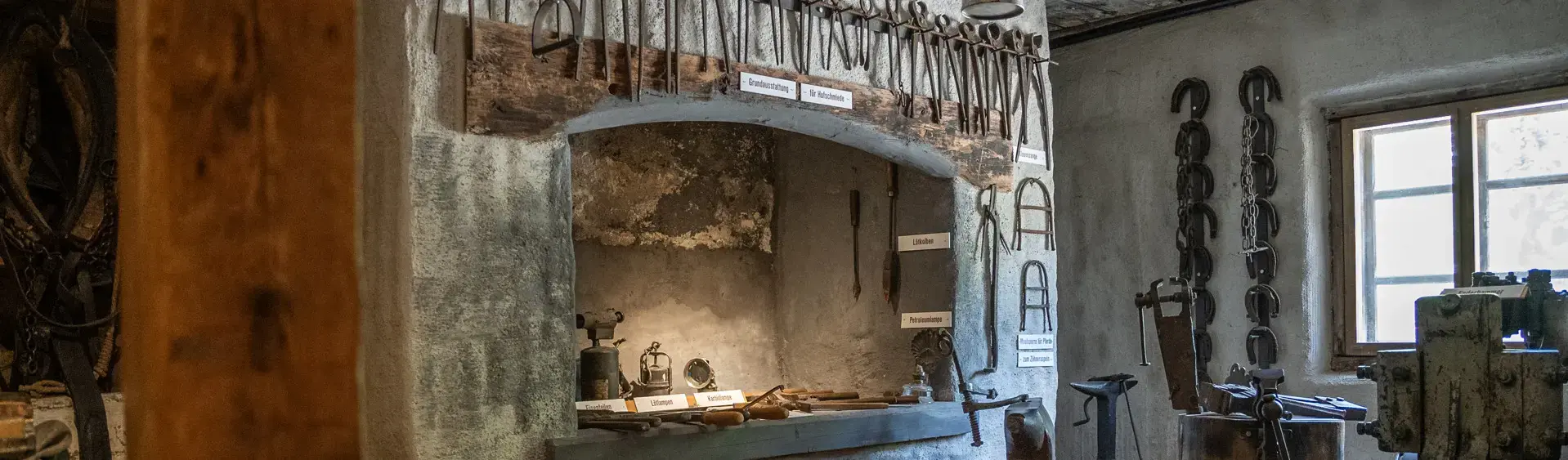 The image depicts an old workshop at the History museum Sixenhof in Achenkirch filled with various tools, including hammers, chisels, and lanterns, organized on a stone wall shelf.