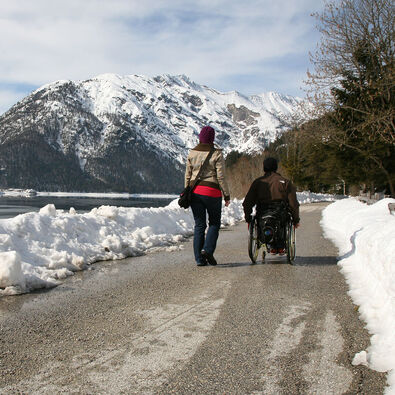 Ein Rollstuhlfahrer und seine Begleiterinn genießen einen Winterspaziergang auf dem Seeuferweg.