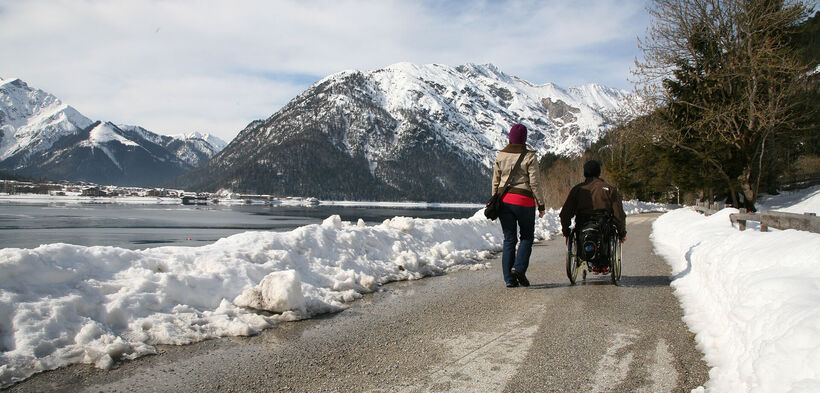 Ein Rollstuhlfahrer und seine Begleiterinn genießen einen Winterspaziergang auf dem Seeuferweg.