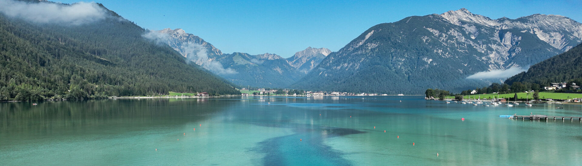 Eine ruhige Landschaft mit dem glitzernden, türkisfarbenen Achensee, umgeben von hohen Bergen und Wäldern. Im Vordergrund wächst das Schilf.
