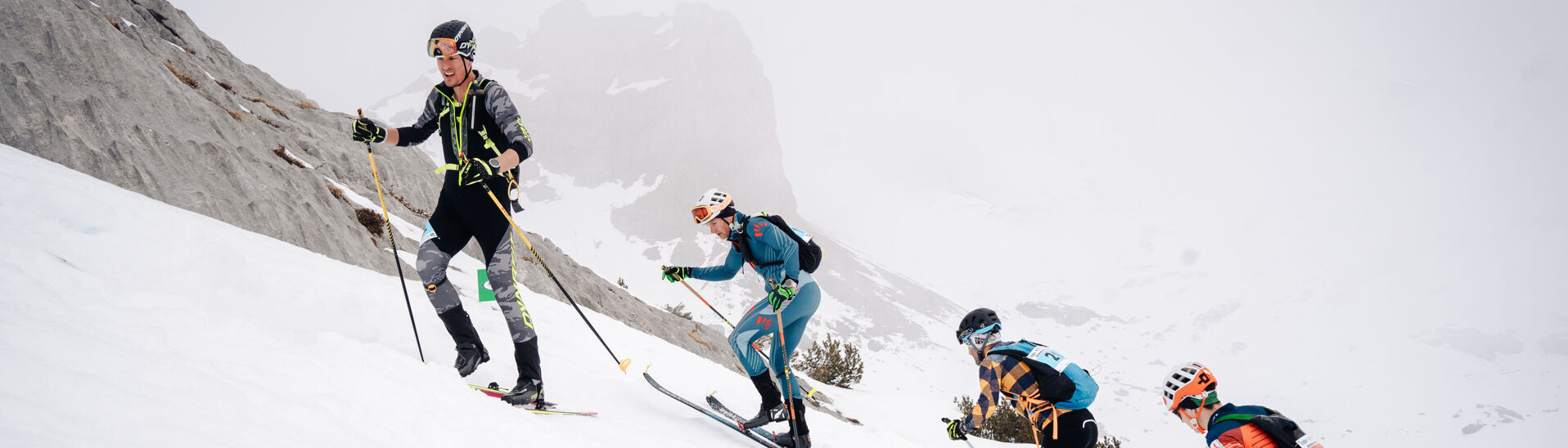 Achensee Xtreme Individual Vier Skifahrer erklimmen einen schneebedeckten Hang im Rofangebirge. Sie tragen technische Ski-Ausrüstung und Skischuhe. Die Umgebung ist gebirgig und in Wolken gehüllt, was die Sicht einschränkt. Alle Sportler sind konzentriert und in Bewegung, während sie den anspruchsvollen Aufstieg bewältigen.