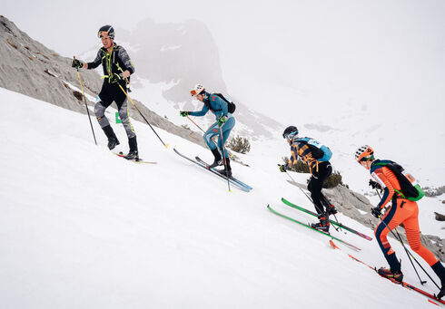 Vier Skifahrer erklimmen einen schneebedeckten Hang im Rofangebirge. Sie tragen technische Ski-Ausrüstung und Skischuhe. Die Umgebung ist gebirgig und in Wolken gehüllt, was die Sicht einschränkt. Alle Sportler sind konzentriert und in Bewegung, während sie den anspruchsvollen Aufstieg bewältigen.