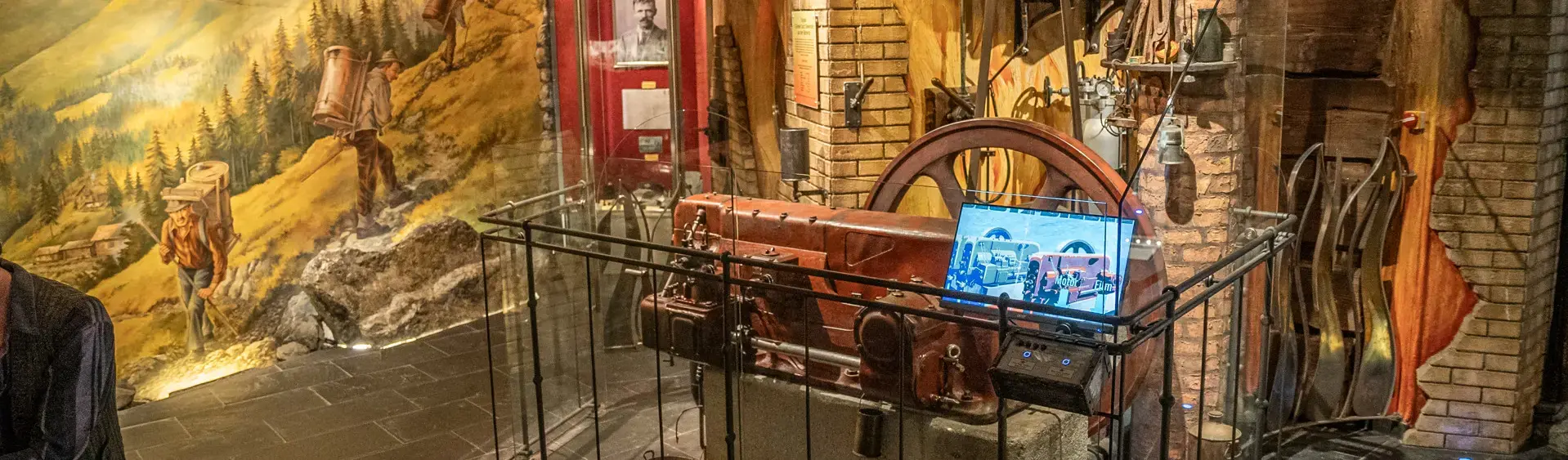 The image displays a historical exhibit featuring a large vintage machine enclosed in glass, with tools and artifacts arranged around it.