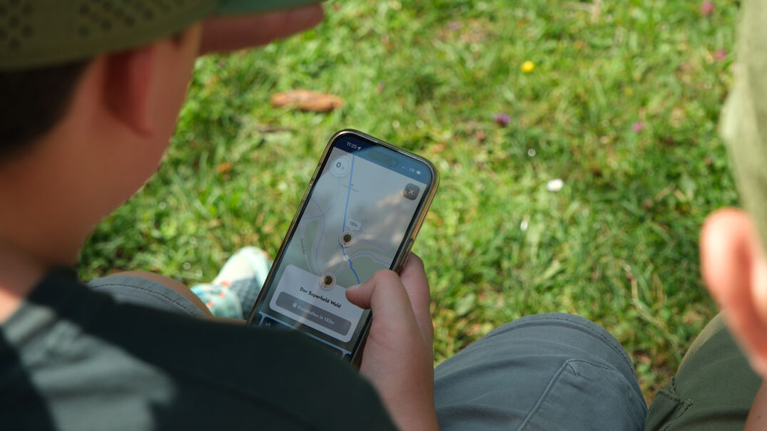 Eine Familie erkundet spielerisch mit dem Handy die wundersame Welt der Fauna und Flora des Naturparks Karwendel auf dem „Achensee Natur Erlebnispfad“.