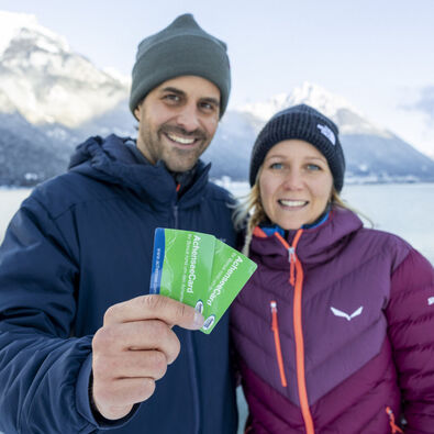 A couple holds the AchenseeCard up to the camera. In the background, the marvellous winter landscape on Lake Achensee.