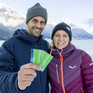 A couple holds the AchenseeCard up to the camera. In the background, the marvellous winter landscape on Lake Achensee.