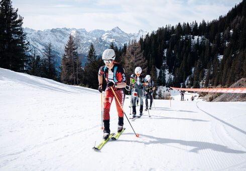 Eine Gruppe von Sportlern steigt auf Skiern einen verschneiten Hang hinauf, umgeben von der beeindruckenden Berglandschaft des Rofangebirges. Sie tragen spezielle Rennanzüge und Helme, während sie die Piste entlangfahren, die von Bäumen und schneebedeckten Bergen umgeben ist.