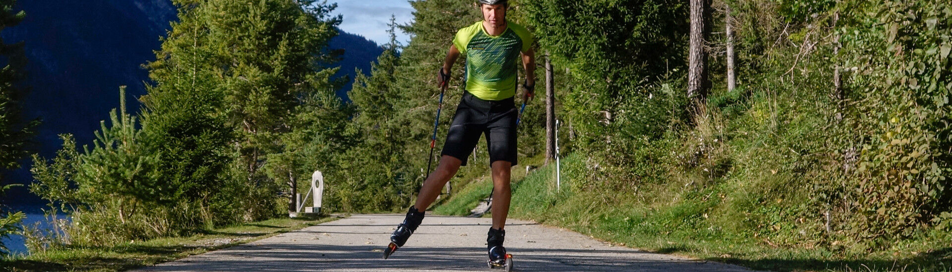 A person wearing a green athletic shirt and black shorts is roller skiing on a paved path at Lake Achensee surrounded by trees and mountains.