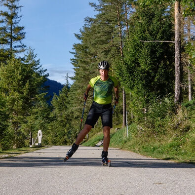 Ein Sportler in einem grünen Trikot fährt auf Skirollern auf einem gepflasterten Weg am Ufer des Achensees entlang.