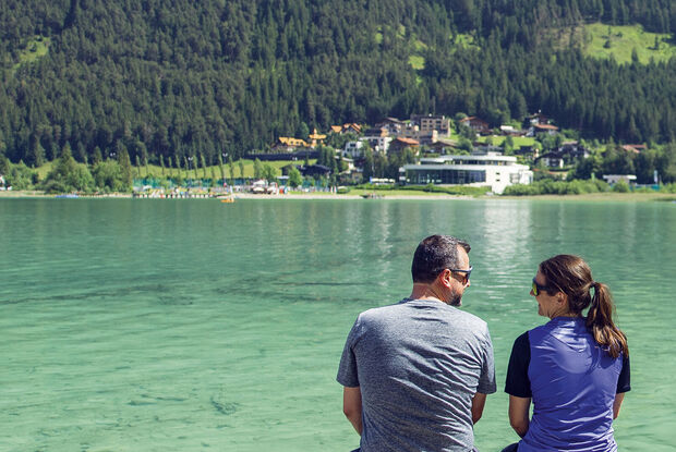 Rast beim Holzsteg in Pertisau am Achensee Ein Paar sitzt am Ufer des Achensees und blickt auf die gegenüberliegende Landschaft von Maurach. Es ist ein sonniger Tag und der Ort vermittelt eine entspannte Atmosphäre.
