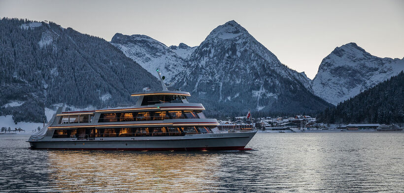 Die MS Achensee bei einer Rundfahrt an einem Winterabend auf dem Achensee. Im Hintergrund sieht man das Dorf Pertisau und das Karwendelgebirge.