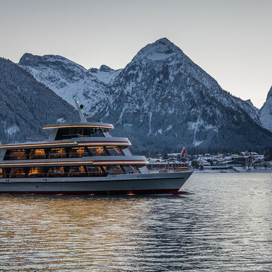 Die MS Achensee bei einer Rundfahrt an einem Winterabend auf dem Achensee. Im Hintergrund sieht man das Dorf Pertisau und das Karwendelgebirge.