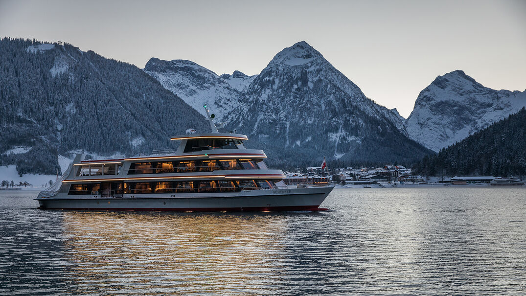Die MS Achensee bei einer Rundfahrt an einem Winterabend auf dem Achensee. Im Hintergrund sieht man das Dorf Pertisau und das Karwendelgebirge.