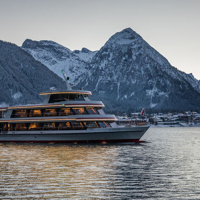 Die MS Achensee bei einer Rundfahrt an einem Winterabend auf dem Achensee. Im Hintergrund sieht man das Dorf Pertisau und das Karwendelgebirge.
