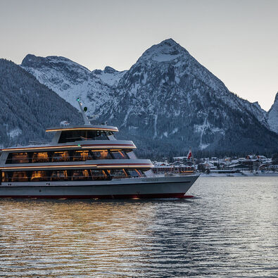 Die MS Achensee bei einer Rundfahrt an einem Winterabend auf dem Achensee. Im Hintergrund sieht man das Dorf Pertisau und das Karwendelgebirge.
