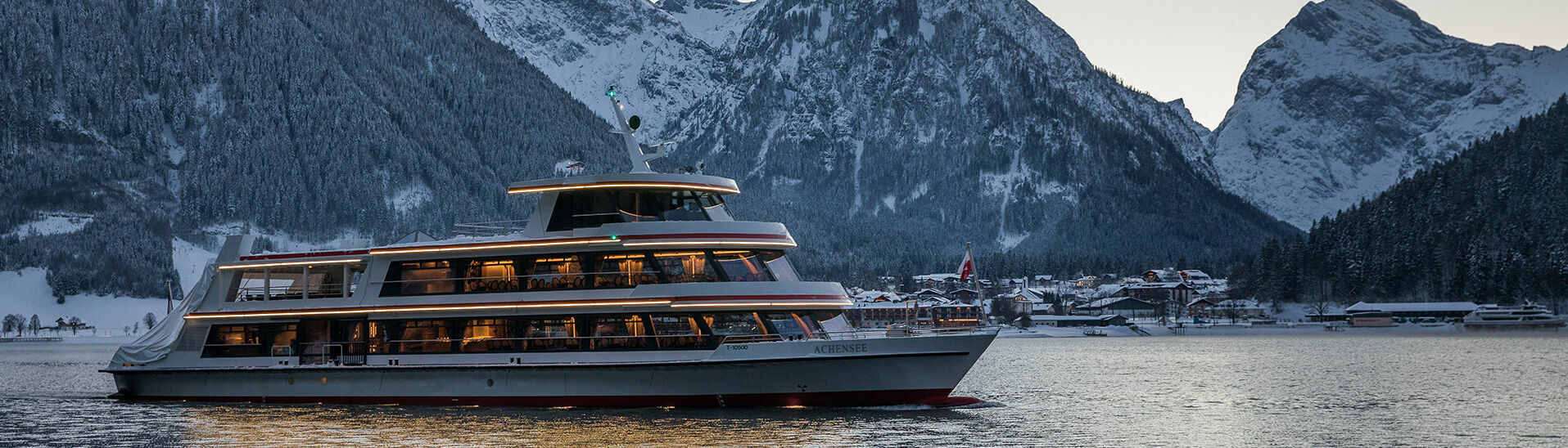 Die MS Achensee bei einer Rundfahrt an einem Winterabend auf dem Achensee. Im Hintergrund sieht man das Dorf Pertisau und das Karwendelgebirge.