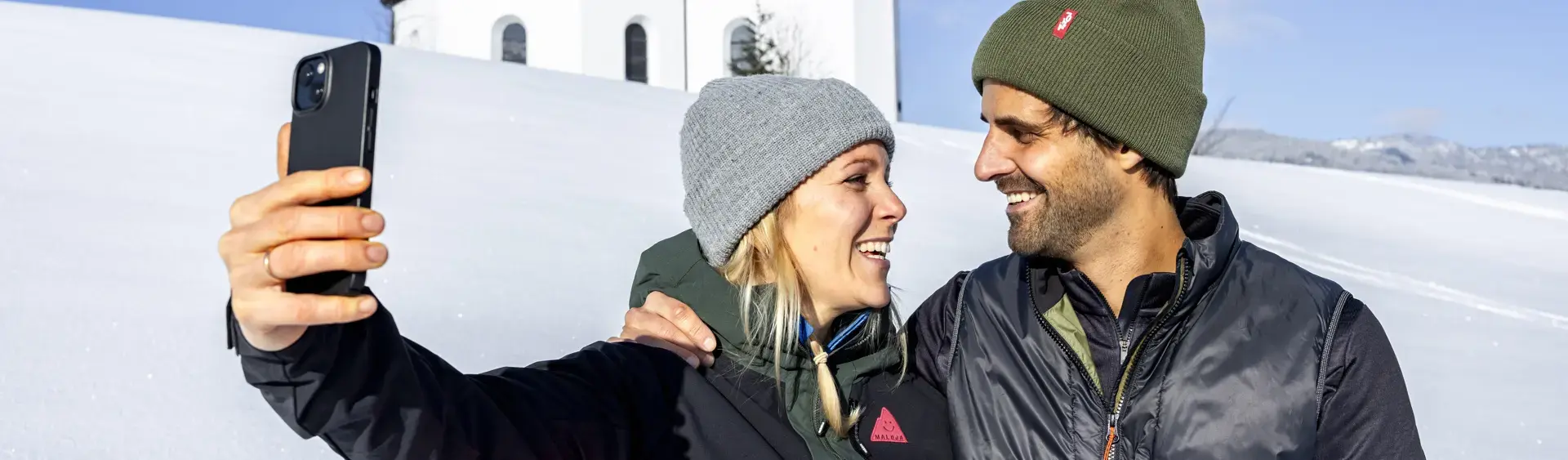 Ein Pärchen möchte den schönen Moment, umgeben von einer herrlichen Winterlandschaft und dem Annakircherl im Hintergrund, mit dem Handy festhalten.