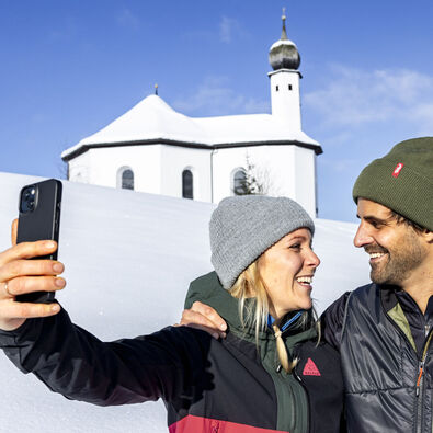 A couple enjoying the beautiful winter landscape near the Annakircherl chapel, capturing the moment on their mobile phone.