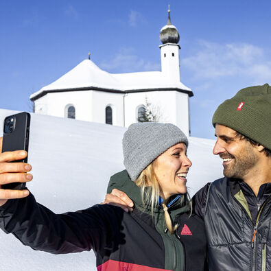 Ein Pärchen möchte den schönen Moment, umgeben von einer herrlichen Winterlandschaft und dem Annakircherl im Hintergrund, mit dem Handy festhalten.
