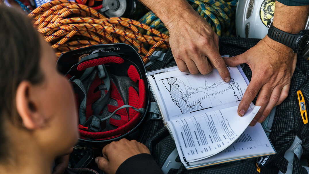 Tourenplanung Ein Mann der einer Frau gerade eine Karte erklärt, welche neben einem Kletterhelm und einem Seil liegt.