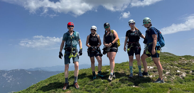 A group of five climbers stands on a grassy peak, wearing helmets and climbing gear. The background features mountains and a clear blue sky with clouds. They appear ready for an outdoor adventure, showcasing a sense of camaraderie and excitement.