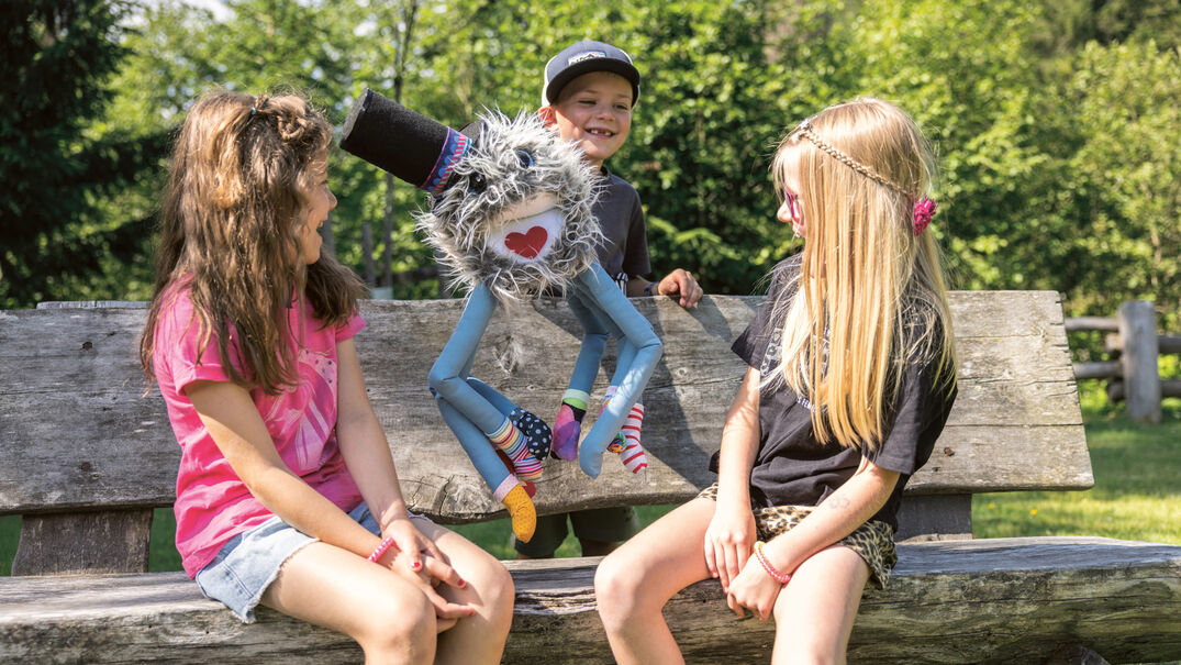 Drei Kinder sitzen auf einer Bank. Ein Junge hält eine farbenfrohe Handpuppe mit einem freundlichen Gesicht und einem fröhlichen Ausdruck. Die Kinder lachen und scheinen Spaß zu haben. Im Hintergrund sind Bäume und grünes Gras zu sehen.