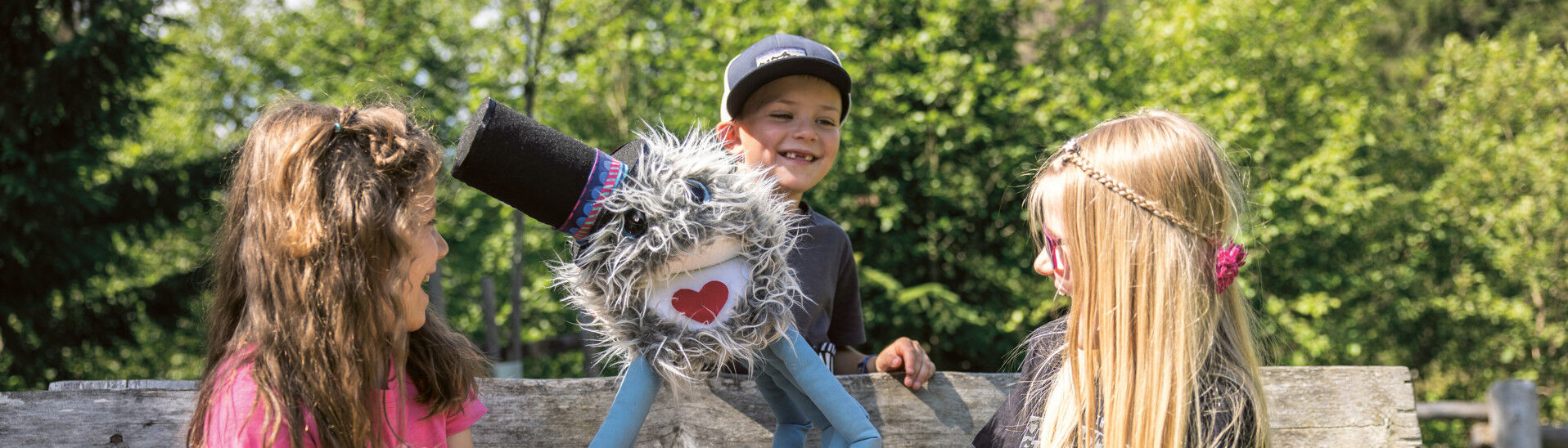 Drei Kinder sitzen auf einer Bank. Ein Junge hält eine farbenfrohe Handpuppe mit einem freundlichen Gesicht und einem fröhlichen Ausdruck. Die Kinder lachen und scheinen Spaß zu haben. Im Hintergrund sind Bäume und grünes Gras zu sehen.