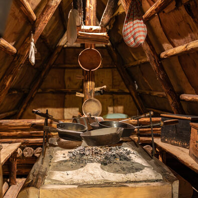 Eine rustikale Küche im Heimatmuseum Sixenhof. In der Mitte steht ein großer Tisch mit einem steinernen Herd, auf dem glühende Kohlen liegen. Darauf sind mehrere Pfannen platziert.
