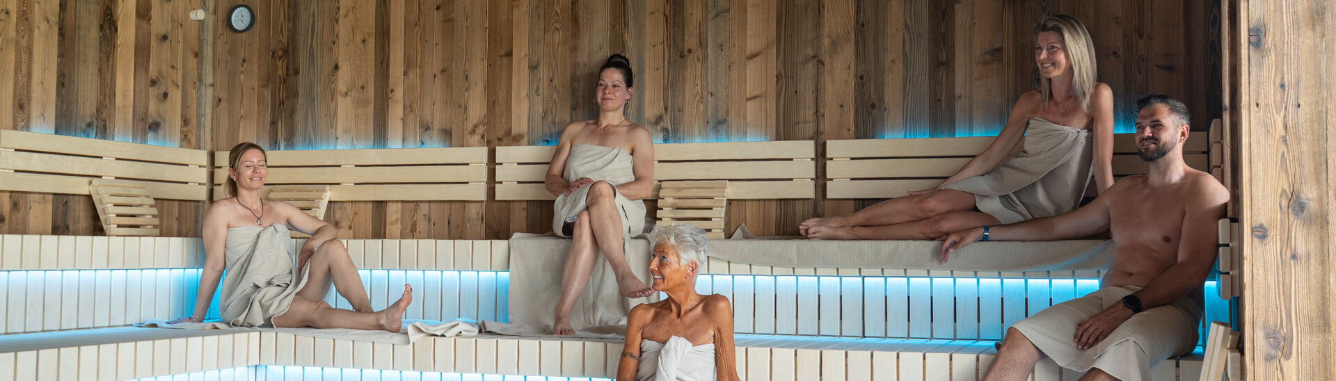 In einer modernen Sauna des Penthouse-SPAs im Atoll Achensee sitzen fünf Menschen in Handtüchern auf Holzbanklinien. Der Raum hat eine warme Atmosphäre mit gedämpftem Licht. In der Mitte ist ein Steingrill sichtbar.