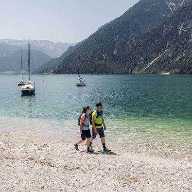 Ein Paar genießt einen kleinen Spaziergang am Ufer des Achensees in Achenkirch. Im Hintergrund sieht man Segelboote.