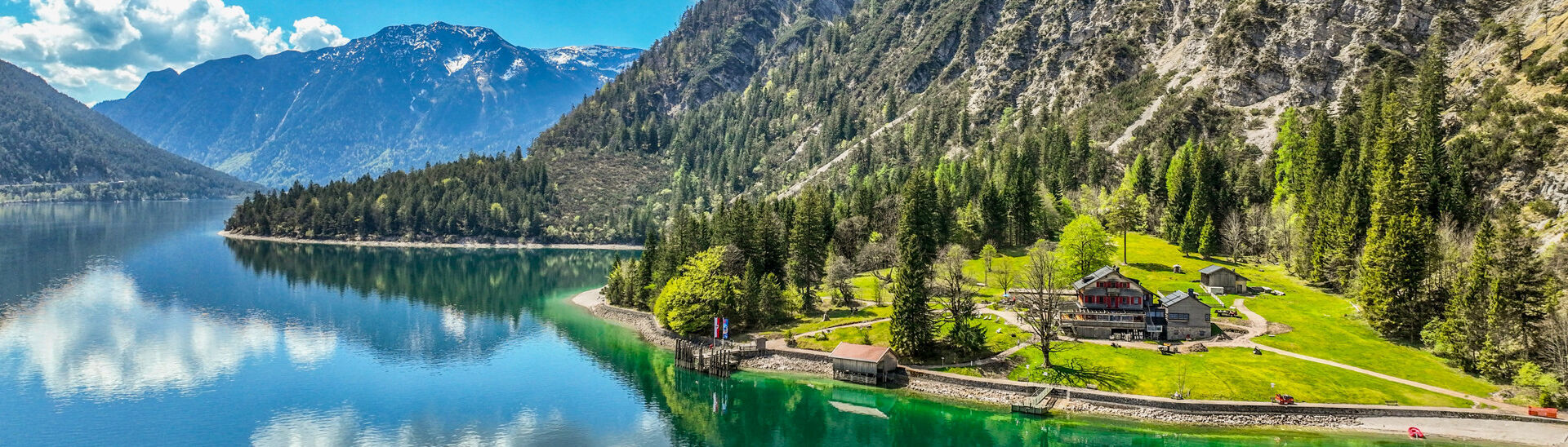 Der Achensee umgeben von hohen Bergen und dichtem Wald. Am Ufer steht die Gaisalm, umgeben von Wiesen. Die blau-weißen Wolken spiegeln sich im Wasser, während die Natur in frischem Grün erblüht.