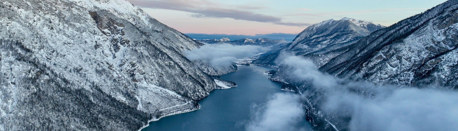 Eine atemberaubende Winterlandschaft zeigt schneebedeckte Berge, die an dem ruhigen, blauen Achensee liegen. Nebelschwaden ziehen über das Wasser und zwischen den Bergen, während der Himmel in sanften Pastellfarben erscheint.