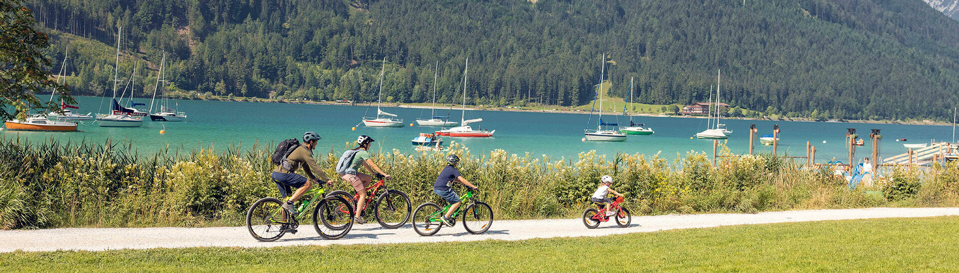 A family with children explores the lakeshore in Maurach am Achensee by bike. In the background of this photo are several sailboats.