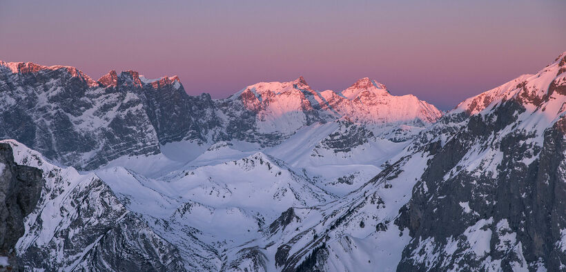 Der Blick auf den winterlichen Naturpark Karwendel bei Sonnenaufgang ist magisch. Die Gipfel und der Himmel färben sich violett und rosarot.