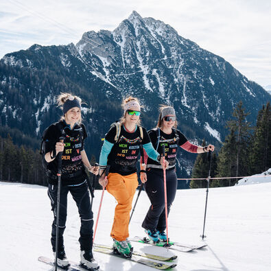 Drei Frauen in Skibekleidung stehen auf einer präparierten Piste im Rofangebirge. Sie lächeln und halten Skistöcke. Im Hintergrund sind majestätische Berge und Bäume sichtbar. Die Stimmung ist fröhlich und sportlich, ideal für einen Skiausflug.