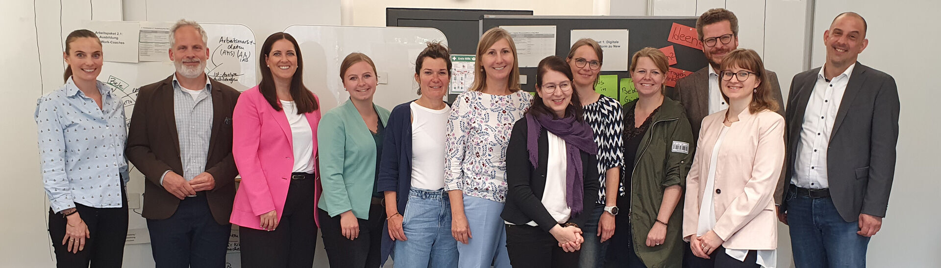 New Work im Tourismus Ein Gruppenfoto von dreizehn New Work Teammitgliedern in einem hellen Raum. Sie lächeln in die Kamera. Im Hintergrund sind Flipcharts und Zeitplanungstafeln sichtbar.