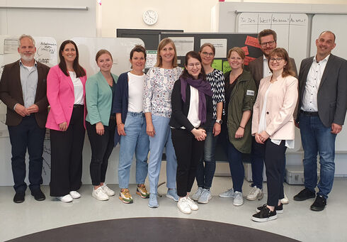 New Work im Tourismus Ein Gruppenfoto von dreizehn New Work Teammitgliedern in einem hellen Raum. Sie lächeln in die Kamera. Im Hintergrund sind Flipcharts und Zeitplanungstafeln sichtbar.