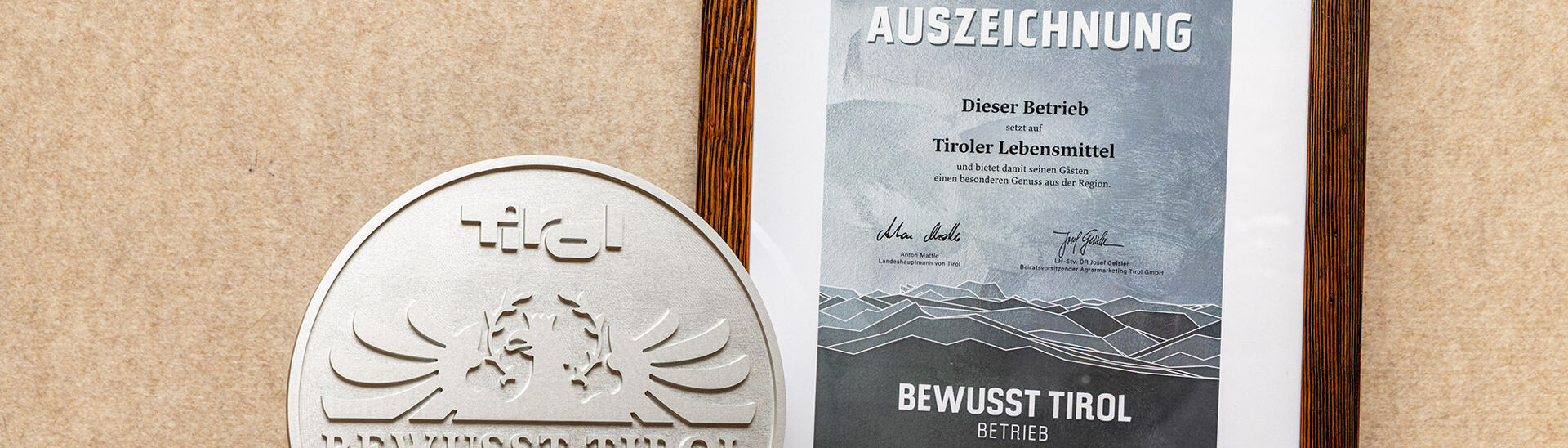 Ein silbernes Qualitätssiegel mit dem Schriftzug "Bewusst Tirol". Daneben ein gerahmtes Zertifikat mit der Aufschrift "2025 AUSZEICHNUNG" und Informationen über die Unterstützung regionaler Lebensmittel.