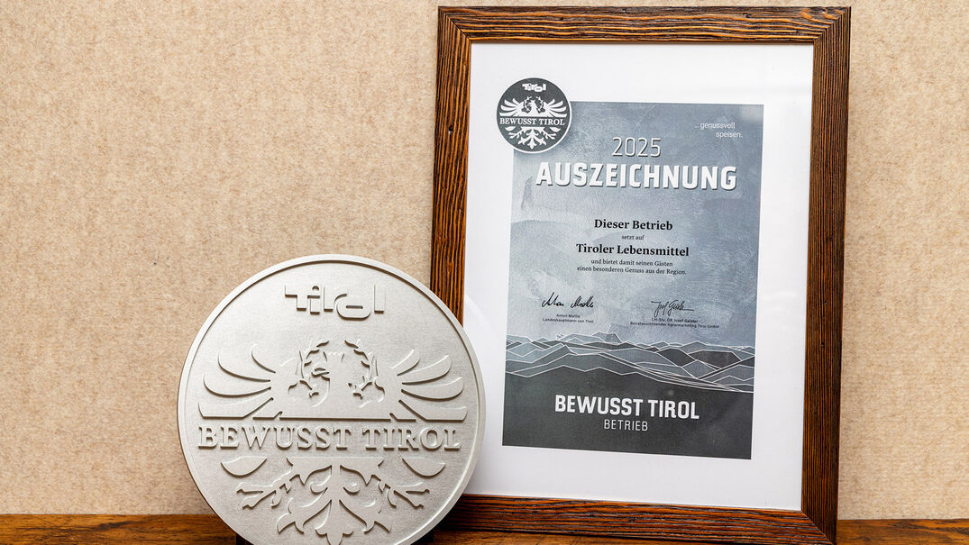 Ein silbernes Qualitätssiegel mit dem Schriftzug "Bewusst Tirol". Daneben ein gerahmtes Zertifikat mit der Aufschrift "2025 AUSZEICHNUNG" und Informationen über die Unterstützung regionaler Lebensmittel.