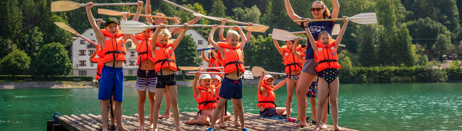 Achensee Kinderprogramm Eine Gruppe von Kindern und einem Erwachsenen steht auf einem Floß im Wasser des Achensees. Sie tragen leuchtend orangefarbene Schwimmwesten und halten Paddel in die Luft. Alle haben fröhliche Gesichtsausdrücke und scheinen Spaß zu haben, während sie gemeinsam posieren.